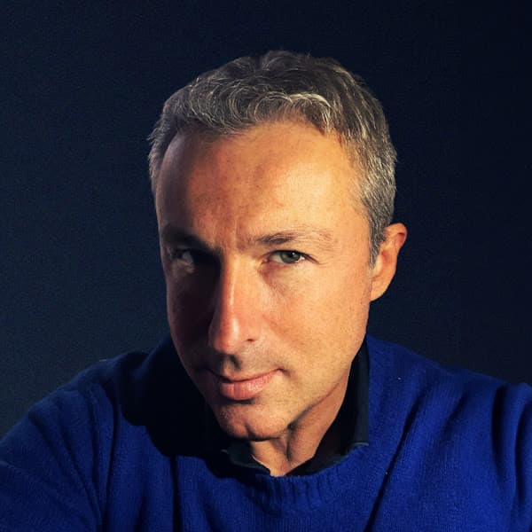 Paolo Chiesa con capelli grigi corti e maglione blu, sguardo diretto, sfondo scuro, illuminazione laterale.